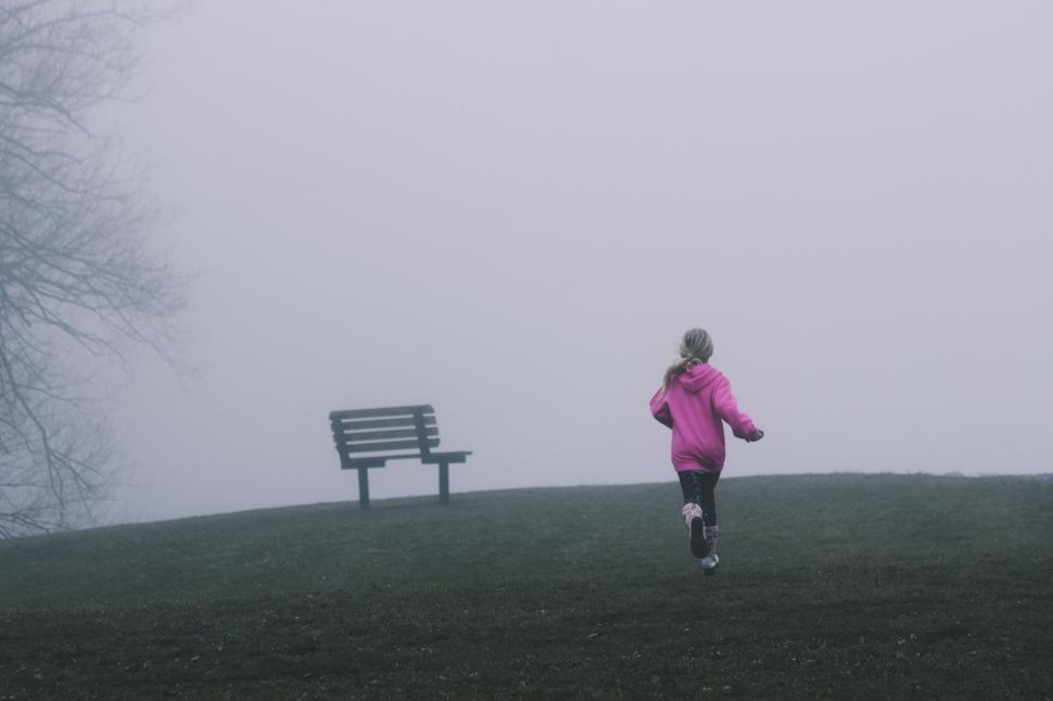 Free Image of Girl running in pink hoodie towards a bench on foggy field. 