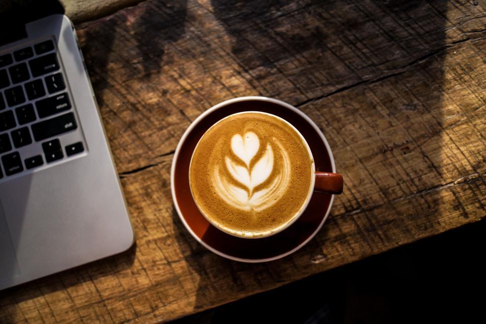 Free Image of Latte art coffee in brown cup beside laptop computer 