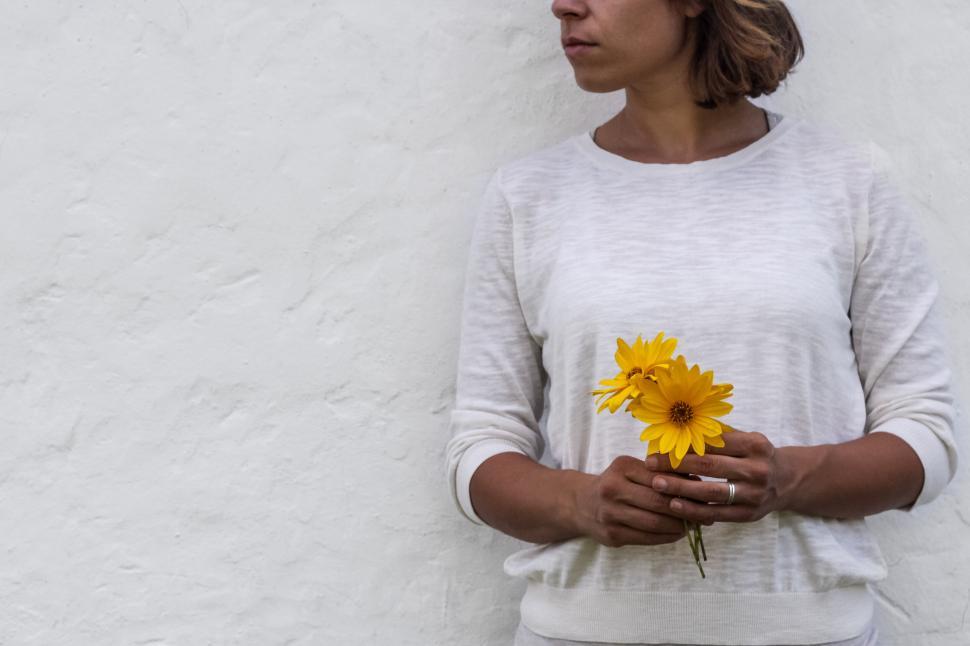 Free Image of Woman holding two yellow flowers against white wall. 