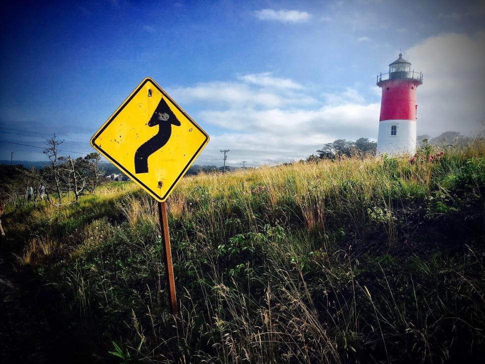 Free Image of Lighthouse next to winding road sign and grassy fields 