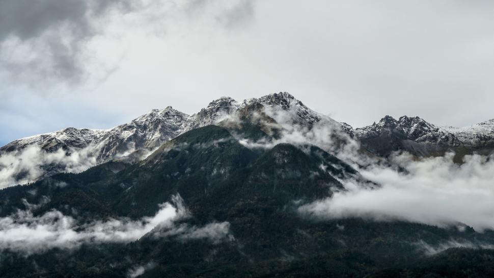 Free Image of Snow-capped mountains with foggy clouds and green trees. 