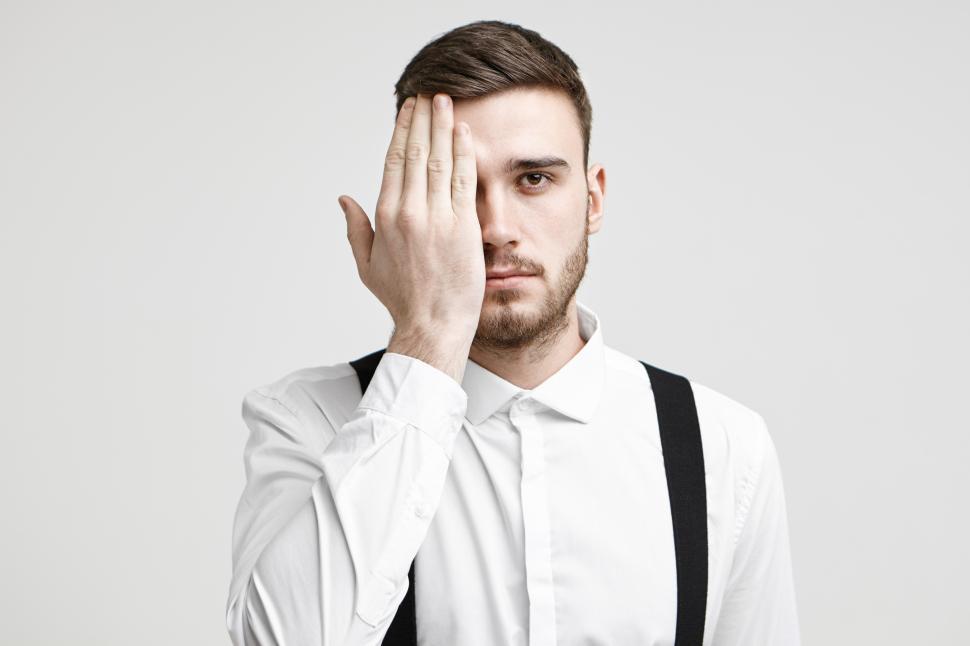 Free Image of Isolated studio shot of handsome young male corporate worker with bristle and stylish haircut looking at camera, covering one eye with palm as if having his eyes tested during vision examination 