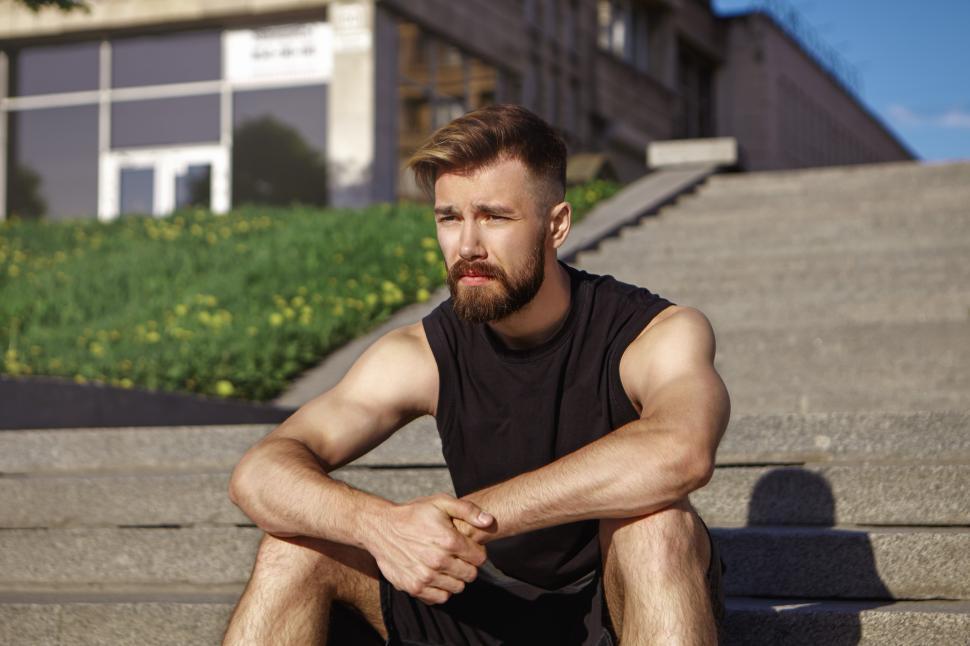 Free Image of Picture of thoughtful exhausted young male runner with fuzzy beard and tanned skin sitting on concrete steps, relaxing after intensive cardio workout Sports, fitness, style and fashion concept 