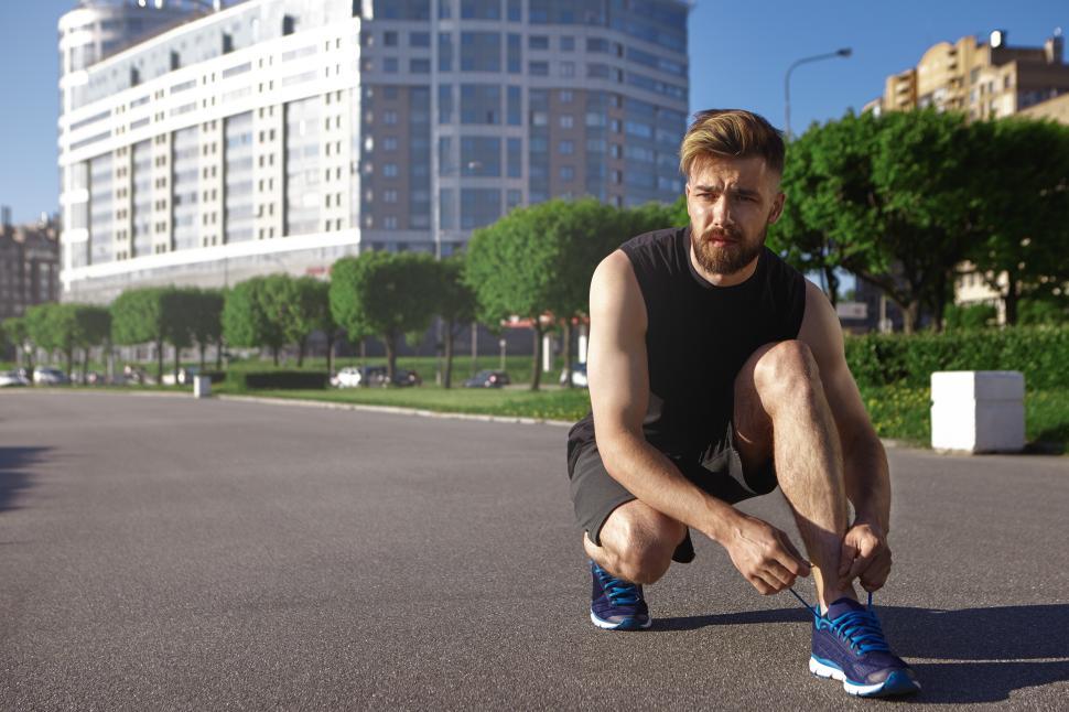 Free Image of Fashionable young unshaven male runner lacing up his stylish running shoes, ready for his morning cardio training. Handsome athletic man in sportswear sitting on road and tying shoes laces 