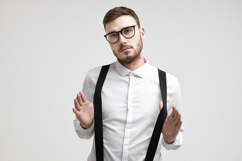 Free Image of Waist up shot of fashionable confident young male office worker with stubble and trendy haircut standing at white wall wearing stylish speactacles and formal shirt, pulling his black suspenders 