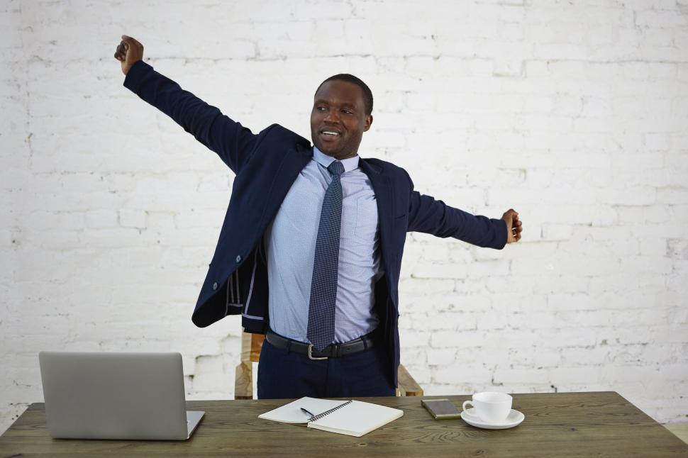 Free Image of Handsome tired dark skinned businessman in elegant suit stretching arms, standing at desk with gadgets, notebook and mug of coffee, feeling relief after he finished work. People, job and occupation 