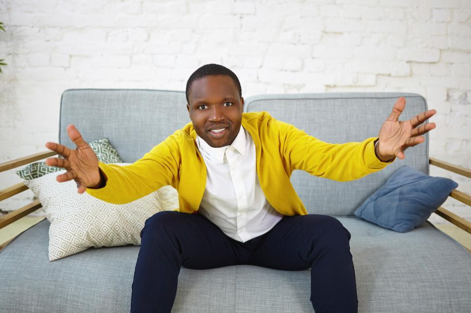 Free Image of Indoor shot of attractive adult dark skinned man sitting on couch in living room and gesturing emotionally, keeping hands wide while watching football game on TV, supporting his favorite team 