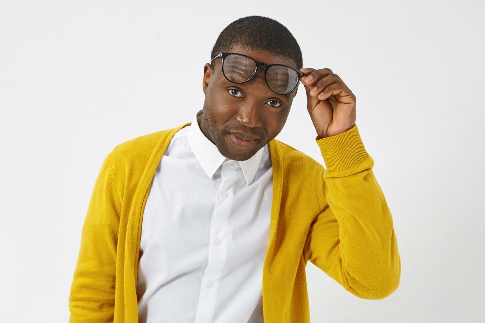 Free Image of People, style, fashion, eyewear, optics and vision concept. Isolated shot of attractive dark skinned guy in stylish wear posing at studio wall, raising trendy spectacles and staring at camera 