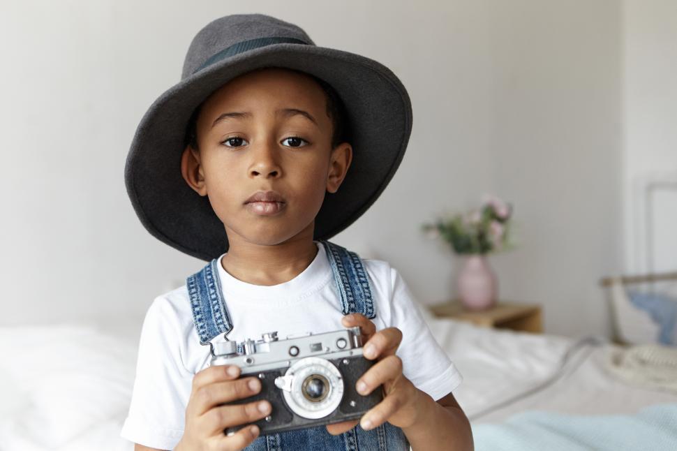 Free Image of People, photography, art and hobby concept. Indoor image of cute adorable Afro American boy posing in cozy bedroom interior wearing stylish round hat, holding vintage camera, taking pictures 