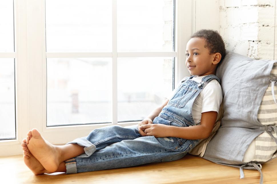 Free Image of Indoor portrait of charming cute eight year old dark skinned African schoolboy wearing jeans jumpsuit spending nice time at home after school, sitting barefooted on windowsill and smiling happily 