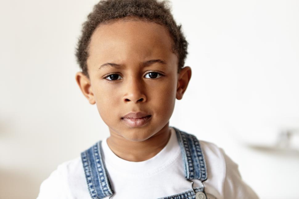 Free Image of Grumpy cute little boy of African appearance dressed in denim jumpsuit and white t-shirt acting naughty, doesn’t want to go to bed, staring at camera, frowning his eyebrows, being in bad mood 