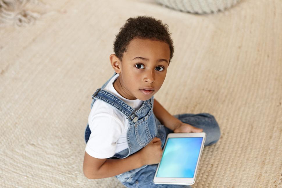 Free Image of Modern gadgets, technology, communication and new generations concept. Top view of cute dark skinned male child sitting on floor and using digital tablet, playing video games online, looking at camera 