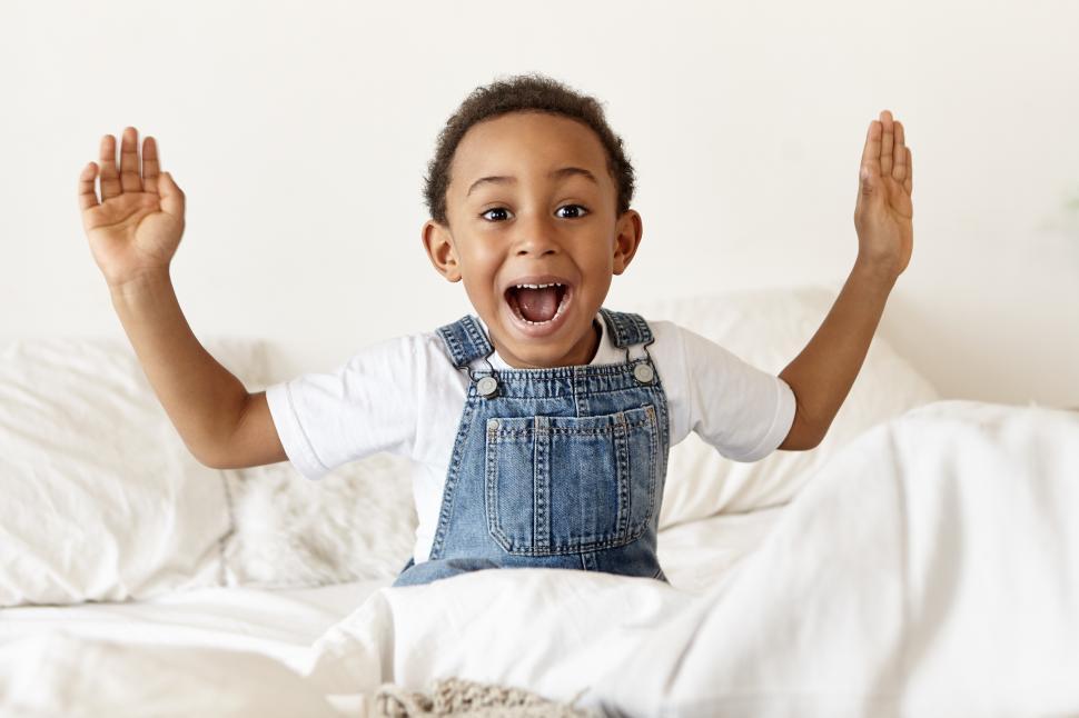 Free Image of Emotional cute little African American boy sitting on white bed, screaming with mouth wide opened and gesturing actively, being in panic because he overslept, late for school. Human emotions 