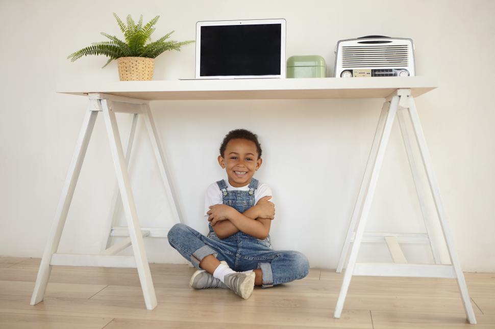 Free Image of Happy childhood, positive human emotions and joy concept. Portrait of adorable cheerful black little boy embracing himself, sitting on floor under white table with laptop and laughing, having fun 