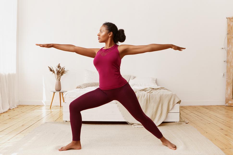 Free Image of Side view of stylish athletic sporty African girl in leggings and top standing barefooted on floor in warrior 2 pose, doing yoga, strengthening legs, training endurance to get strong healthy body 