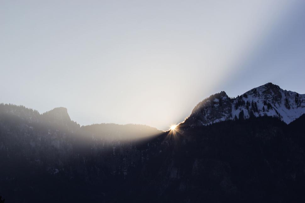 Free Image of Sunlight peeking over a mountain with snow-capped peaks 