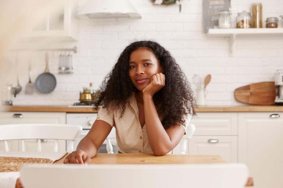 Free Image of Positive beautiful young African American female with voluminous hair sitting in kitchen at table using portrable computer. Social media, networking, technology and modern lifestyle concept 