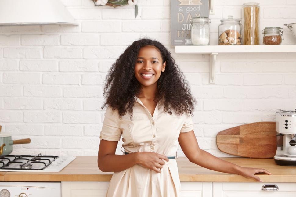Free Image of Food, cuisine, cookery and household concept. Beautiful cheerful young dark-skinned woman with Afro hairstyle standing at kitchen counter, making breakfast for her family, smiling broadly at camera 