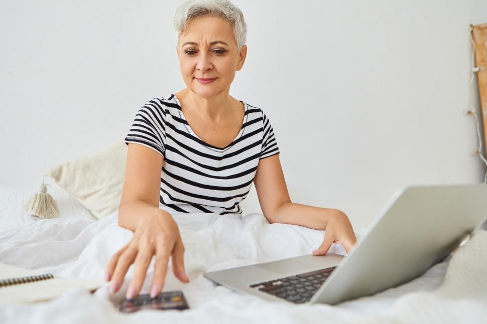 Free Image of Attractive gray haired elderly businesswoman working remotely right from bedroom, sitting on bed with portable computer, using calculator, managing finances, having confident happy expression 