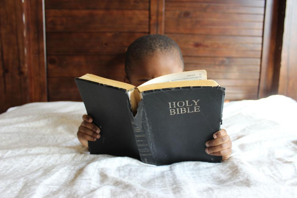 Free Image of Child reading Holy Bible in bed with hands holding book. 