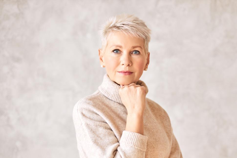Free Image of Serious pensive middle aged female wearing warm pullover and pearl earrings looking at camera having deep in thoughts facial expression, holding hand under chin. Contemplation and deliberation 