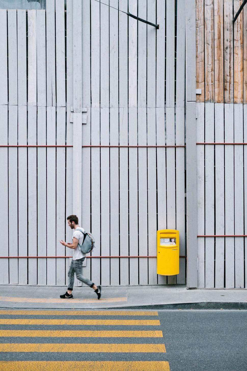 Free Image of Man with backpack walking along a street with fence. 