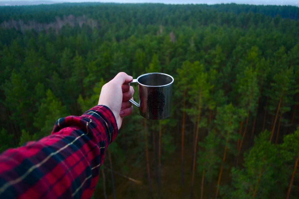 Free Image of Hand holding metal cup above a vast dense forest landscape 