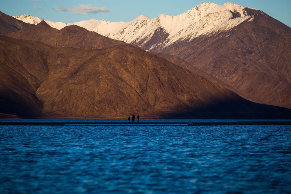 Free Image of Distant hikers near a mountain lake with snow peaks 