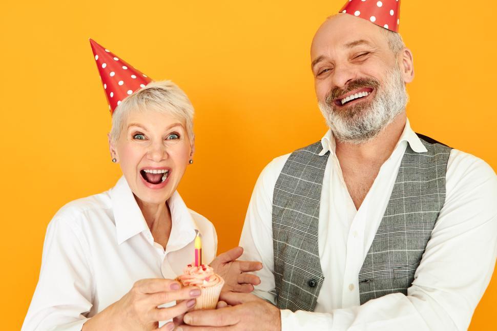 Free Image of Love, family, celebration, joy and happiness. Beautiful overjoyed short haired mature female celebrating wedding anniversary with her bearded husband, wearing cone hats and blowing candle on cup cake 