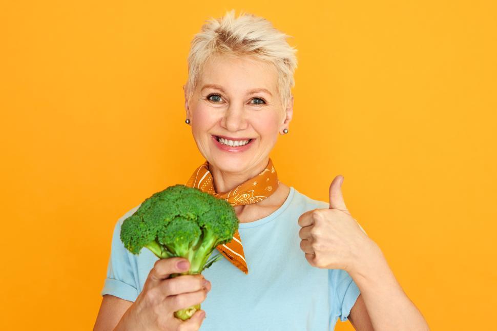 Free Image of Attractive middle aged woman with short pixie hair holding fresh broccoli and smiling at camera choosing healthy organic food. Mature female making raw meal using vegetables, showing thumbs up 