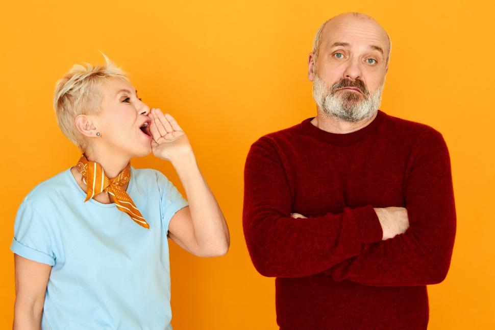 Free Image of Annoyed mature female with short blonde hair holding hand at her mouth shouting something at husband who is ignoring, not listening to her, keeping arms folded on his chest. Relationships concept 