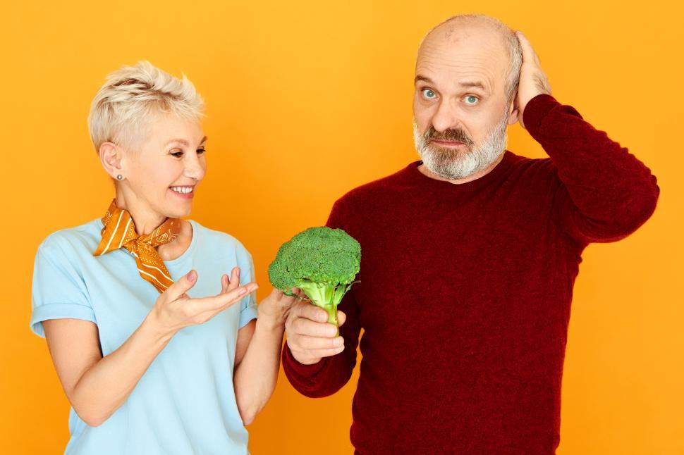 Free Image of Dieting, food, health, organic products and vegetarianism concept. Frustrated senior man looking at camera with mournful facial expression, holding disgusting broccoli, his wife makes him eat greens 