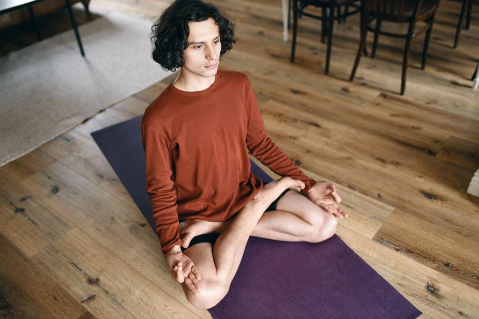 Free Image of High angle view of focused young man with flexible body sitting in lotus posture on mat, meditating with eyes open, havinf mindful look, concentrating on some object, relaxing body, slowing down 