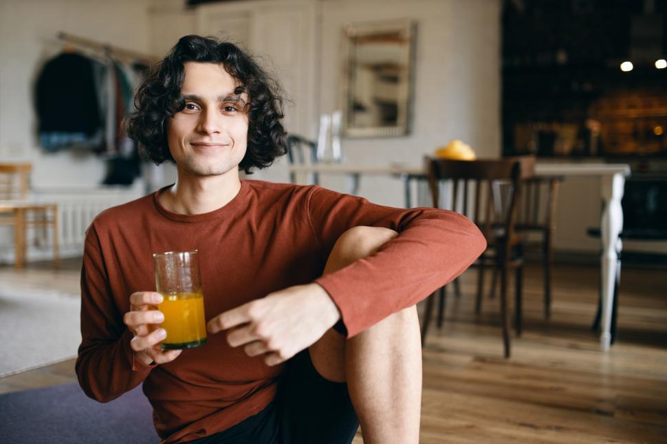 Free Image of Healthy lifestyle, eating, food, nutrition and dieting concept. Cheerful handsome young man sitting on floor, having orange fresh for breakfast being on juice fasting, smiling happily at camera 