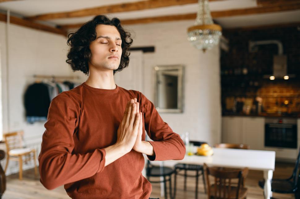 Free Image of Portrait of handsome young man with black curly hair posing in cozy kitchen interior keeping eyes closed and pressing hands together in namaste, praying, doing meditation, having calm look 