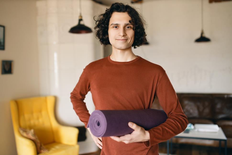 Free Image of Positive young man in casual clothes holding roll of yoga mat under his arm going to practice indoors, happy to stay home during quarantine, spending more time on self development and healthy activity 
