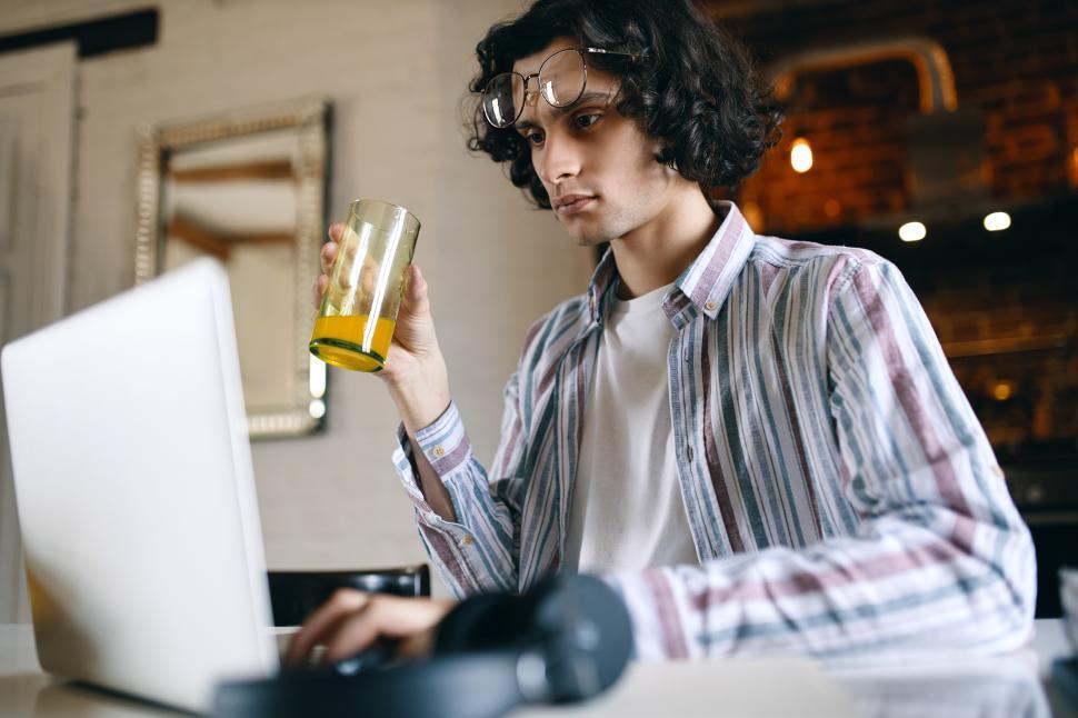Free Image of Concentrated serious young businessman in casual shirt working from home, sitting at desk in front of open laptop, checking email in the morning, drinking healthy fresh orange juice for breakfast 
