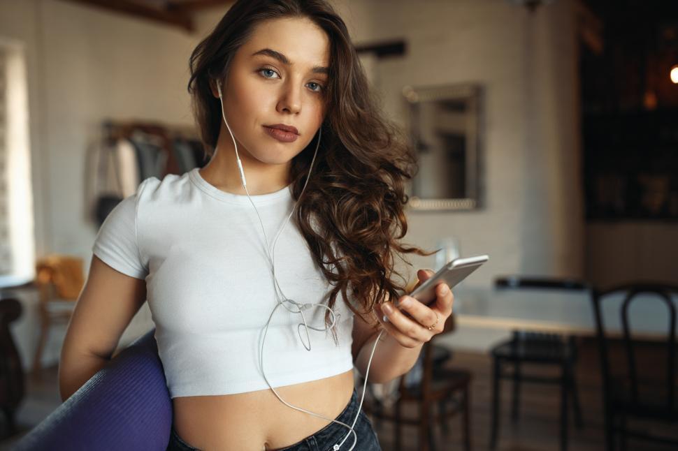 Free Image of Portrait of chubby attractive teenage girl with long wavy hair and make up posing at home using earphones, messaging online using mobile phone, holding fitness mat, staying always connected 