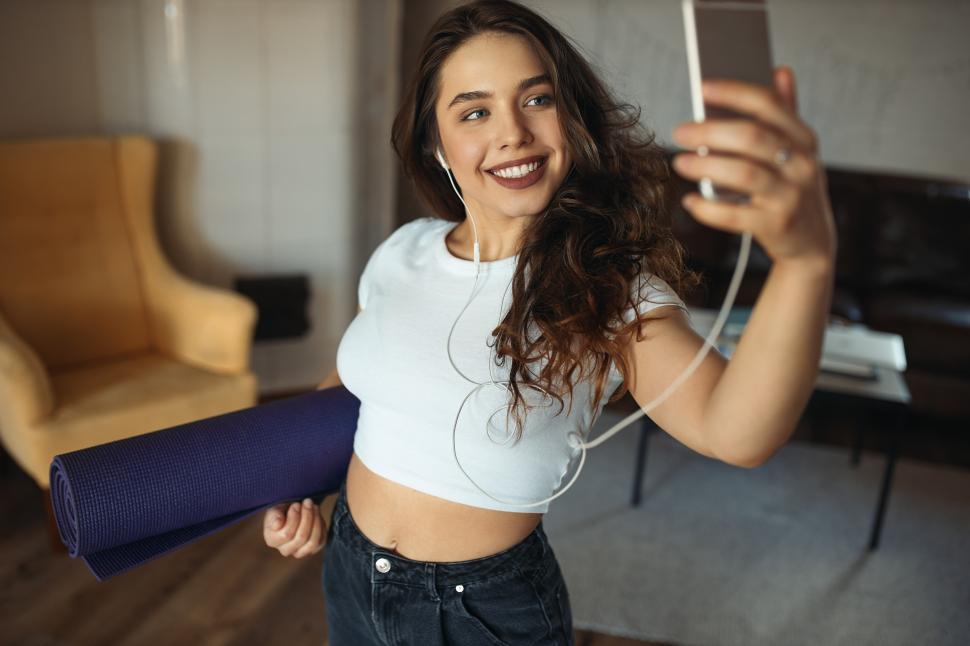 Free Image of Indoor image of cute charming teenage girl in crop top and jeans holding yoga mat and smart phone, posing for selfie in earphones, smiling happily. Active lifestyle, training and modern gadgets 