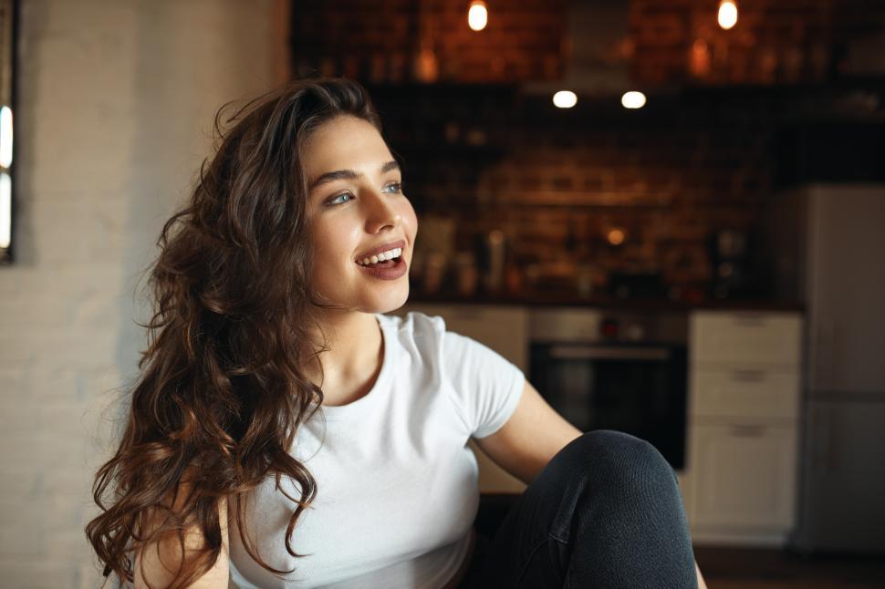 Free Image of Horizontal shot of pretty girl with long curly hair and blue eyes sitting comfortably at kitchen table opening mouth wide, singing, being in good mood, having fun, looking with dreamy happy smile 