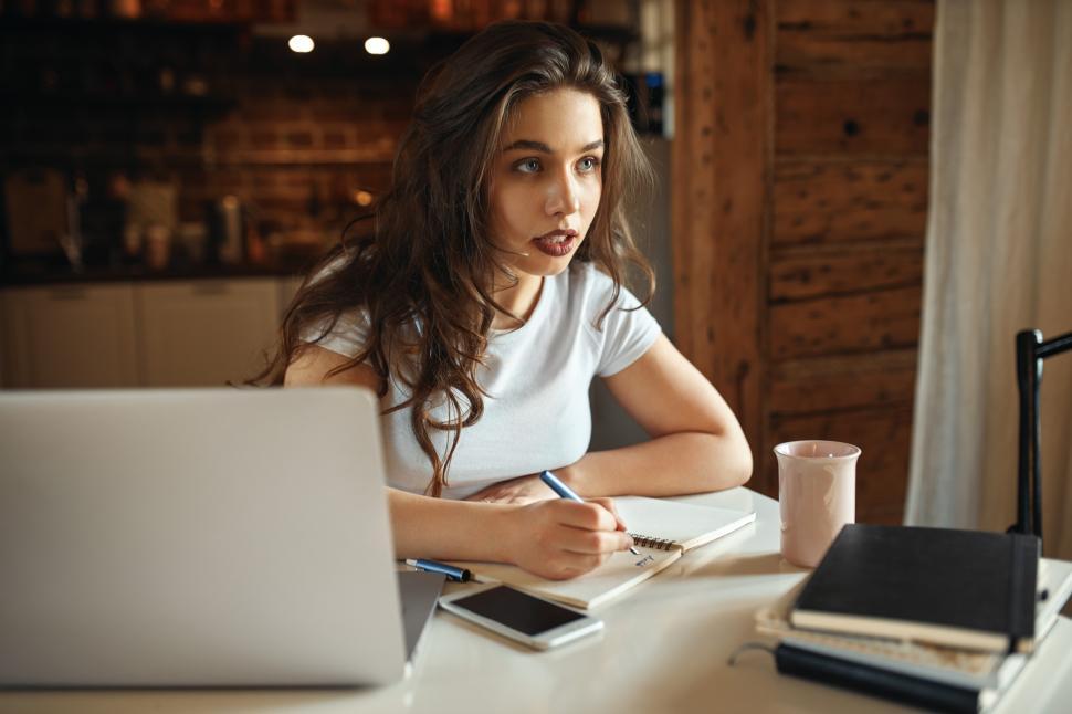 Free Image of Pretty stylish stuent girl holding pen making notes in notebook doing homework, writing essay, sitting at table with textbooks and portable computer, having thoughtful serious facial expression 