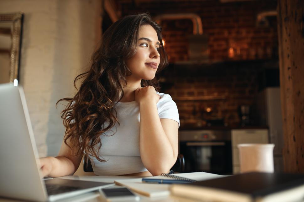 Free Image of Cute young female with long wavy hair enjoying sunny day using portable computer for distant work, having dreamy look. Charming student girl studying online on laptop at home. Selective focus 