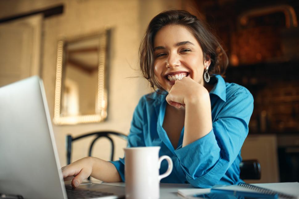 Free Image of Cheerful attractive young woman enjoying distant work, sitting at desk using portable computer, drinking coffee. Pretty female blogger working from home, uploading video on her channel, smiling 