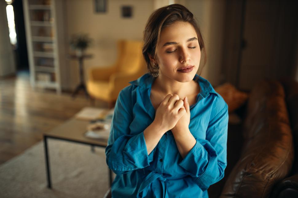 Free Image of Portrait of joyful charming young female sitting on sofa in living room closing eyes, keeping hands on her chest, praying, having calm peaceful facial expression, dreaming of something pleasant 