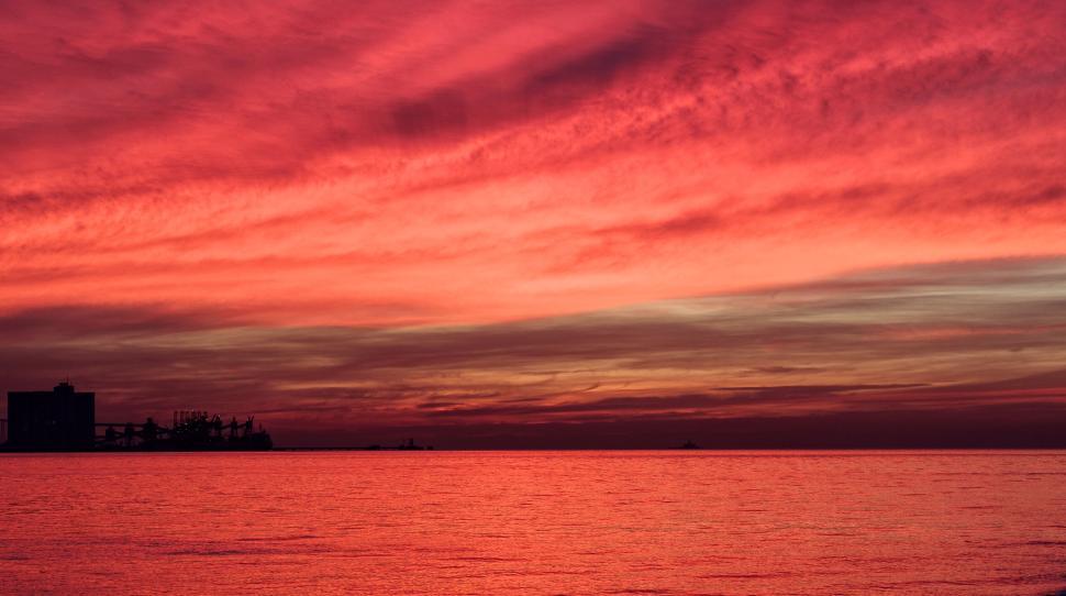 Free Image of Red and pink sunset over calm ocean with silhouette shore 