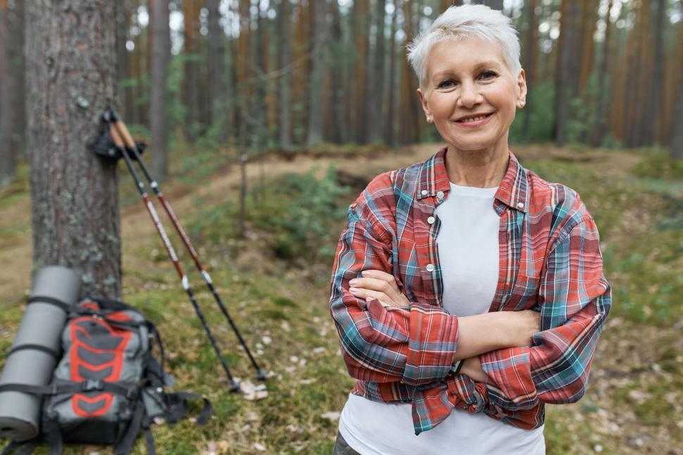Free Image of Energetic attractive middle aged woman hiker posing outdoors, choosing active lifestyle, traveling alone with backpack and nordic poles for walking, keeping arms crossed and smiling at camera 