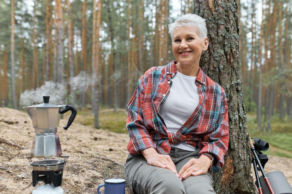 Free Image of Camping lifestyle in forest. Cheerful middle aged Euroepan woman sitting on ground under pine going to make tea, boiling water in kettle on gas stove burner, having joyful happy facial expression 