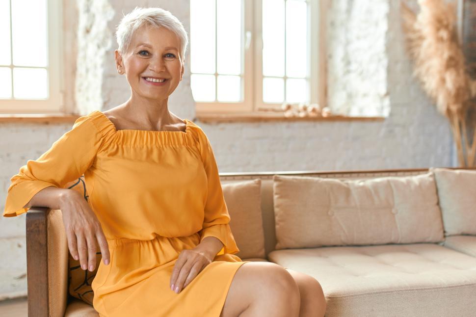 Free Image of People, lifestyle, leisure, retirement and relaxation. Indoor shot of beautiful fashionable European female pensioner in yellow dress sitting comfortably on couch in living room, smiling happily 