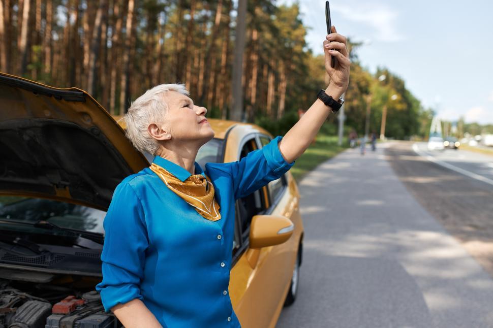 Free Image of Outdoor image of unhappy middle aged woman standing on road by broken car with open hood raising hand with mobile phone, looking for network signal, trying to call for assistance. No connection 