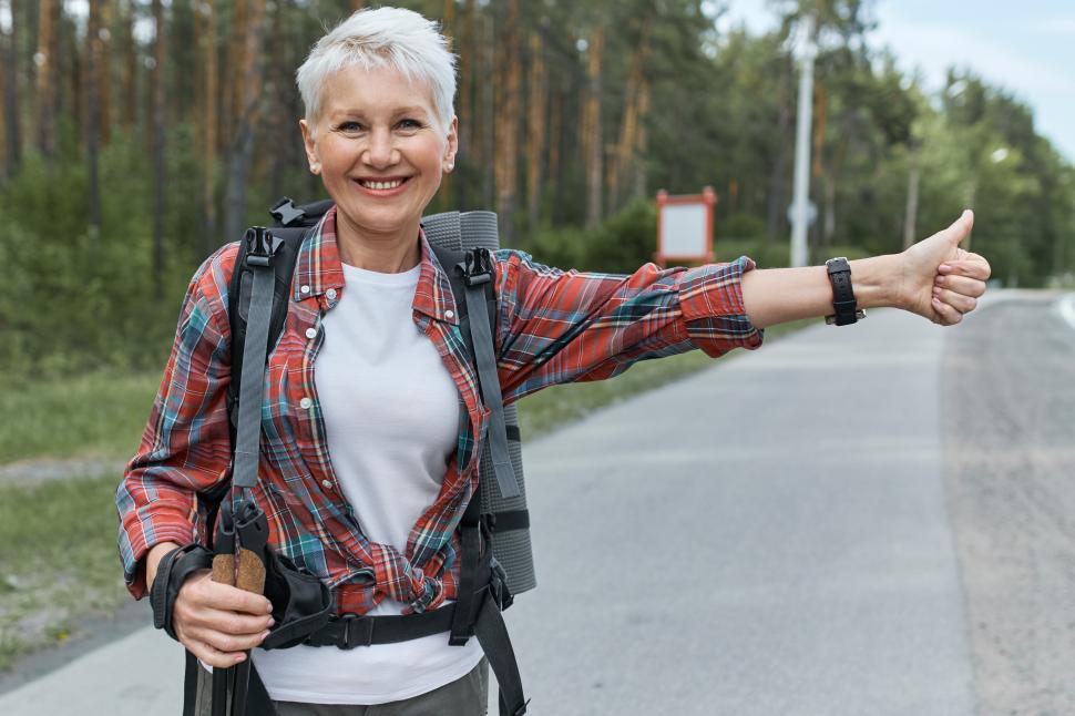 Free Image of Energetic female pesnioner in active wear standing on road with rucksack behind her back hitchhiking making sign with thumbs up gesture, signaling that she needs a ride. Travel and vacations 